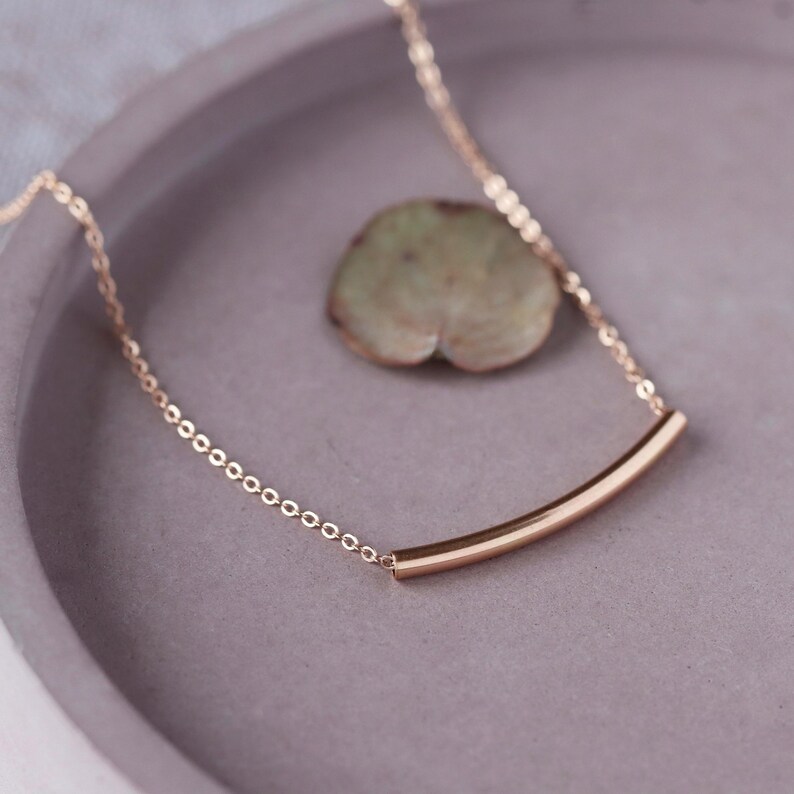 May include: A delicate gold-colored necklace with a curved bar pendant. The necklace chain is made of small, connected links. The pendant is a smooth, curved bar. The necklace rests on a gray surface with a green leaf.