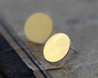 May include: A pair of gold-colored, circular stud earrings. The earrings have a brushed metal finish and are set against a vintage paper background. The earrings are simple and minimalist.