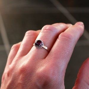 May include: A silver ring with a dark red gemstone. The gemstone is round and set in a decorative crown-like setting. The ring band is thin and simple.