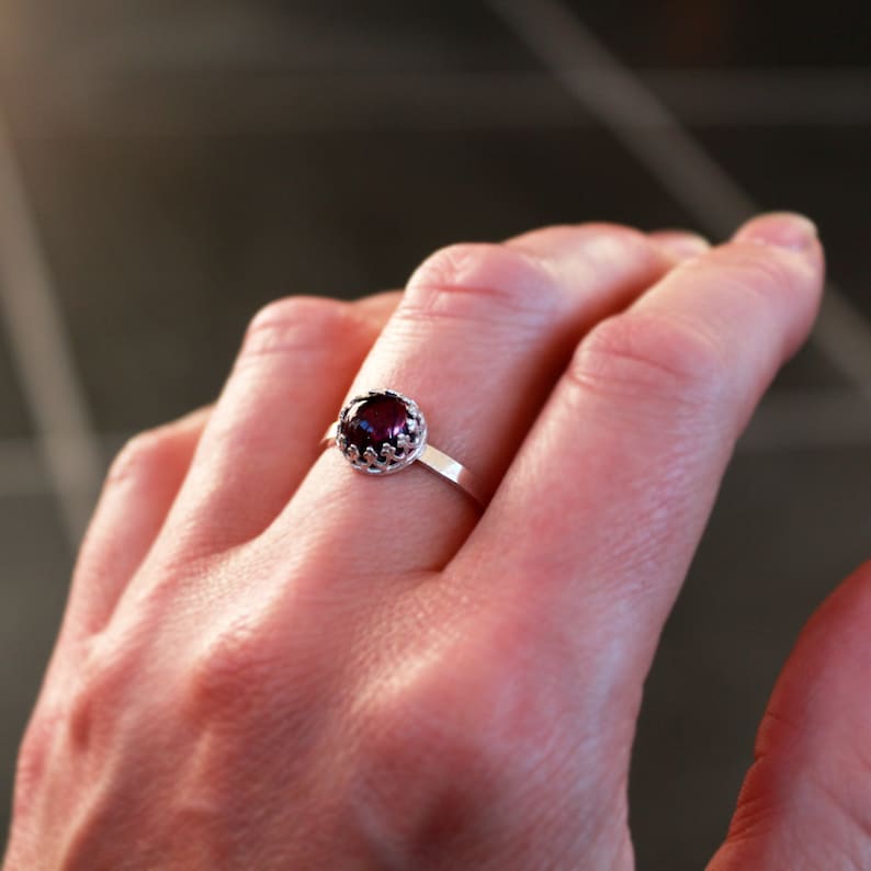 May include: A silver ring with a dark red gemstone. The gemstone is round and set in a decorative crown-like setting. The ring band is thin and simple.