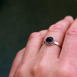 May include: A silver ring with a large, round, dark purple gemstone. The gemstone is set in a decorative, openwork bezel. The ring is on a finger, with a blurred background.