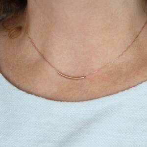 May include: A delicate rose gold necklace featuring a curved bar pendant. The pendant is suspended from a fine chain, creating a minimalist and elegant design. The necklace is shown against a light background.