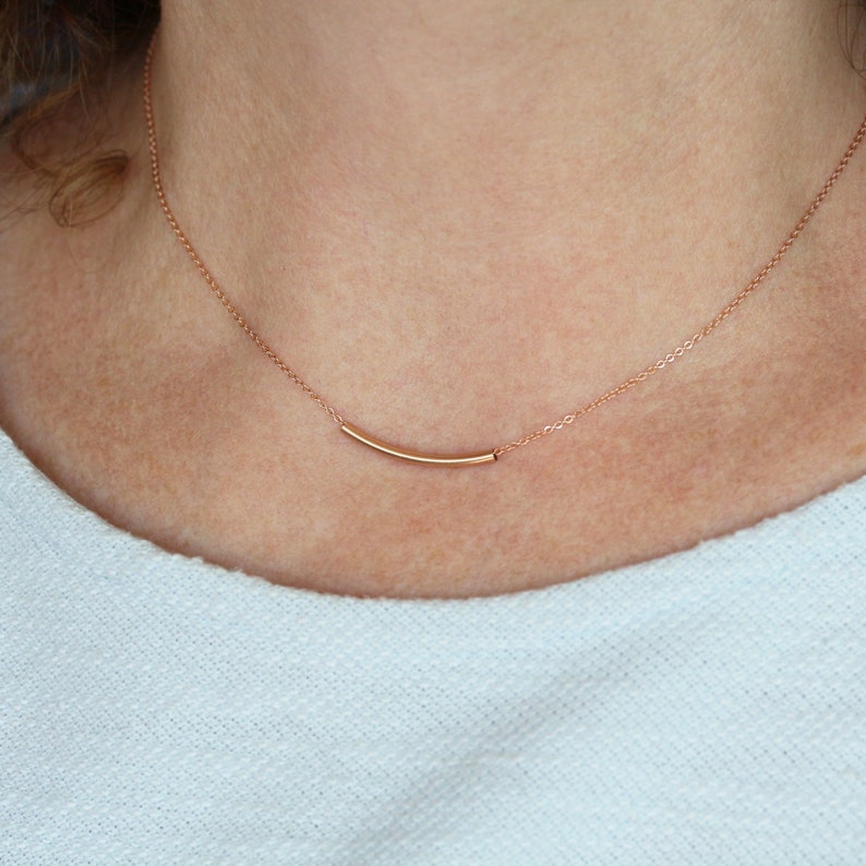 May include: A delicate rose gold necklace featuring a curved bar pendant. The pendant is suspended from a fine chain, creating a minimalist and elegant design. The necklace is shown against a light background.