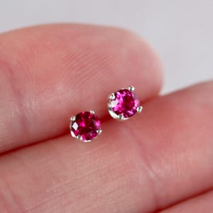 May include: A pair of stud earrings featuring vibrant, round, magenta-colored gemstones set in silver-colored metal. The gemstones have multiple facets, reflecting light. The earrings are small and delicate, suitable for everyday wear.