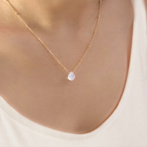 May include: Delicate gold necklace featuring a small, pear-shaped, iridescent moonstone pendant. The necklace has a fine chain with tiny gold beads spaced along its length. The moonstone reflects a soft, milky white color, adding a touch of elegance.