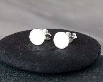 May include: Two silver stud earrings with a circular, white face. The earrings are displayed on a dark, textured surface. The earrings are simple and minimalist, suitable for everyday wear.
