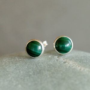 May include: A pair of round stud earrings featuring polished, vibrant green malachite stones set in silver-colored bezels. The earrings are displayed on a light gray surface, showcasing their natural color and design.
