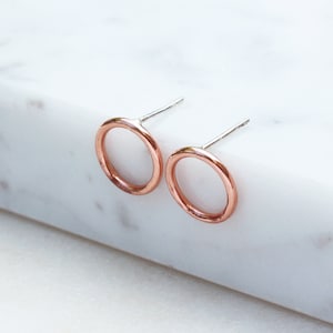 May include: A pair of open circle stud earrings. The earrings are made of copper-colored metal with silver posts. The earrings are displayed on a white marble surface.