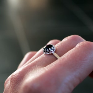 May include: A silver ring with a dark, oval-shaped gemstone. The gemstone is set in a decorative, openwork crown setting. The ring is on a finger, with a blurred background.
