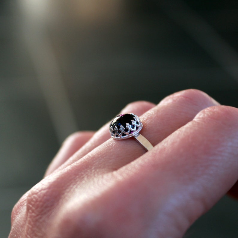 May include: A silver ring with a dark, oval-shaped gemstone. The gemstone is set in a decorative, openwork crown setting. The ring is on a finger, with a blurred background.
