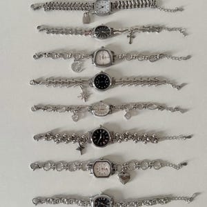 Könnte beinhalten: Eine Sammlung von acht silberfarbenen Armbanduhren mit verschiedenen Zifferblattformen und Gliederarmbändern. Einige Uhren haben Anhänger wie ein Schloss, ein Kreuz, einen Mond, einen Stern und ein Herz. Die Zifferblätter sind weiß oder schwarz.