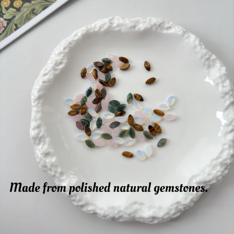 May include: A white, textured ceramic dish holds an assortment of polished gemstones in shades of brown, pink, green, and white. The gemstones are oval-shaped. Text at the bottom reads: Made from polished natural gemstones.