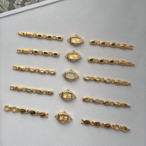 May include: A collection of gold-toned vintage watches and watch bands. The watches have rectangular and oval faces with gold hands. The bands feature various colored stones and decorative elements. The pieces are arranged on a light-colored surface.