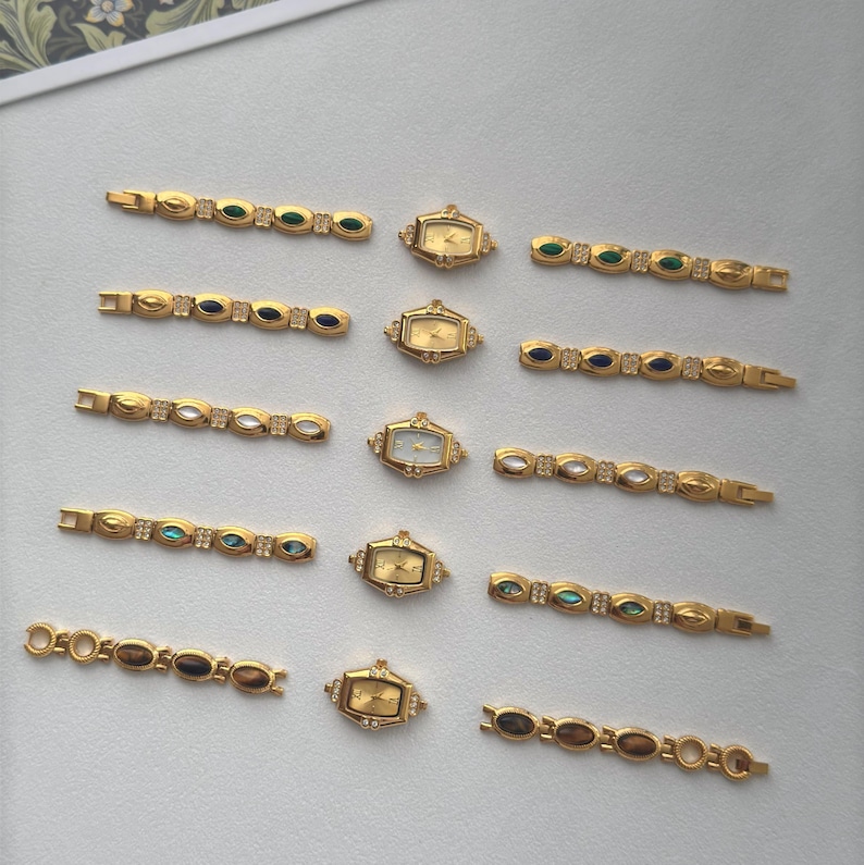 May include: A collection of gold-toned vintage watches and watch bands. The watches have rectangular and oval faces with gold hands. The bands feature various colored stones and decorative elements. The pieces are arranged on a light-colored surface.