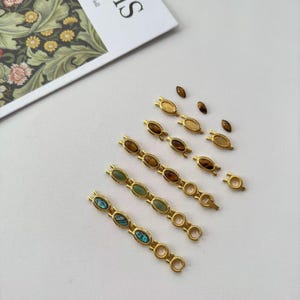May include: Gold-toned jewelry components with oval-shaped gemstones in various colors, including brown, green, and iridescent blue. The components are arranged on a white surface, suggesting a craft or jewelry-making theme.
