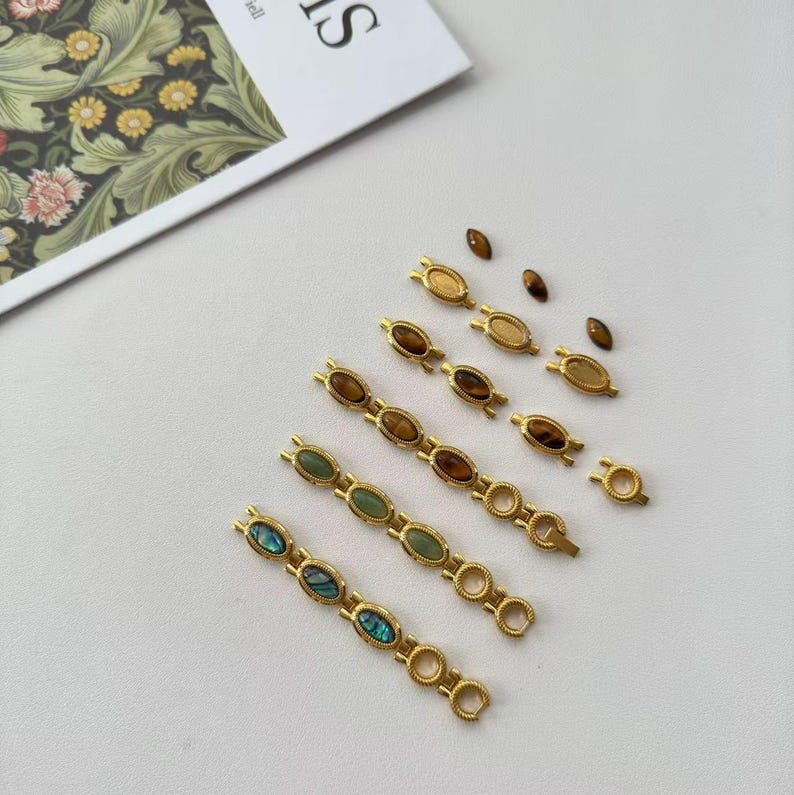 May include: Gold-toned jewelry components with oval-shaped gemstones in various colors, including brown, green, and iridescent blue. The components are arranged on a white surface, suggesting a craft or jewelry-making theme.