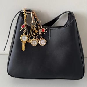 Puede incluir: Bolso de hombro negro con herrajes dorados y un charm decorativo. El charm presenta una llave, un corazón y un sol, cada uno con una pequeña esfera de reloj. El bolso tiene forma curva y una sola correa para el hombro.
