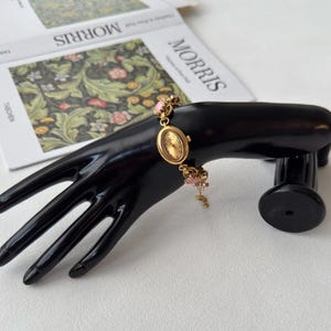 Pu&ograve; includere: Un orologio da polso dorato con quadrante ovale e una delicata catena impreziosita da dettagli rosa. L'orologio &egrave; esposto su un supporto nero a forma di mano, con un libro intitolato "MORRIS" sullo sfondo.