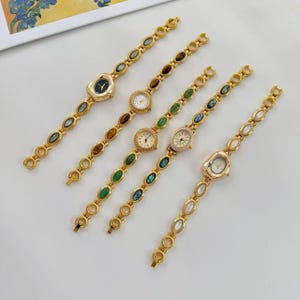 May include: Five gold-toned wristwatches with decorative bands. Each watch features a different face and gemstone accents in various colors, including green, brown, and iridescent blue. The watch faces are round or oval, with delicate hands and markers.