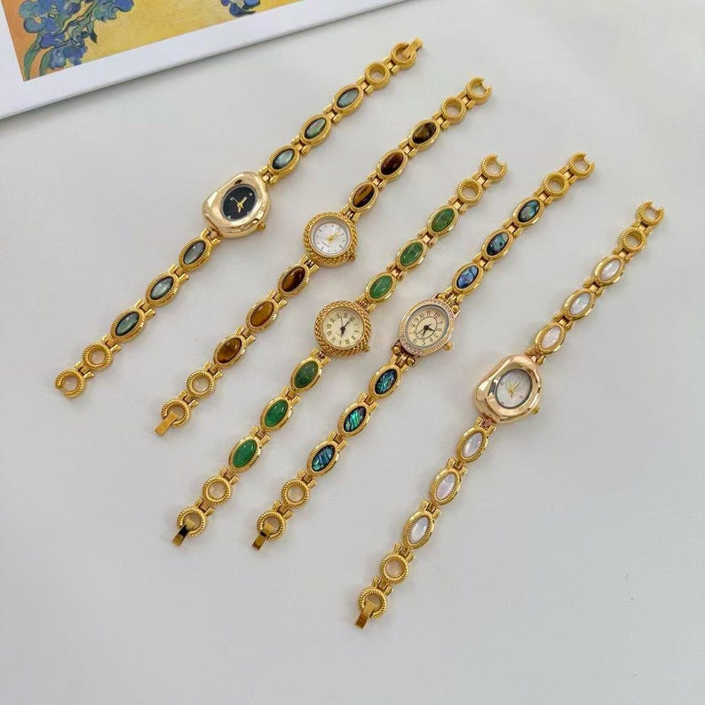 May include: Five gold-toned wristwatches with decorative bands. Each watch features a different face and gemstone accents in various colors, including green, brown, and iridescent blue. The watch faces are round or oval, with delicate hands and markers.