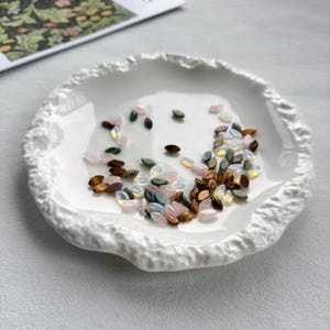 K&ouml;nnte beinhalten: Eine wei&szlig;e, strukturierte Keramikschale enth&auml;lt eine Auswahl an kleinen, ovalen Perlen. Die Perlen sind in den Farbt&ouml;nen Braun, Rosa, Gr&uuml;n und schillerndem Wei&szlig; gehalten. Die Schale hat einen wellenf&ouml;rmigen, organischen Rand.