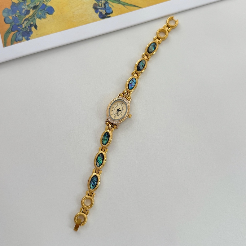 May include: Gold-toned wristwatch with an oval face and a band adorned with abalone shell accents. The watch face has a cream-colored background with black numerals and hands. The band features oval abalone shell insets, creating a vintage aesthetic.