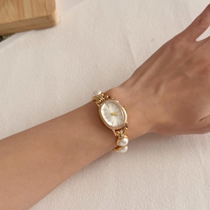 Puede incluir: Un reloj de pulsera dorado con una esfera ovalada y blanca. La correa del reloj presenta perlas decorativas. El reloj se lleva en la mu&ntilde;eca, mostrando su elegante dise&ntilde;o y estilo cl&aacute;sico.