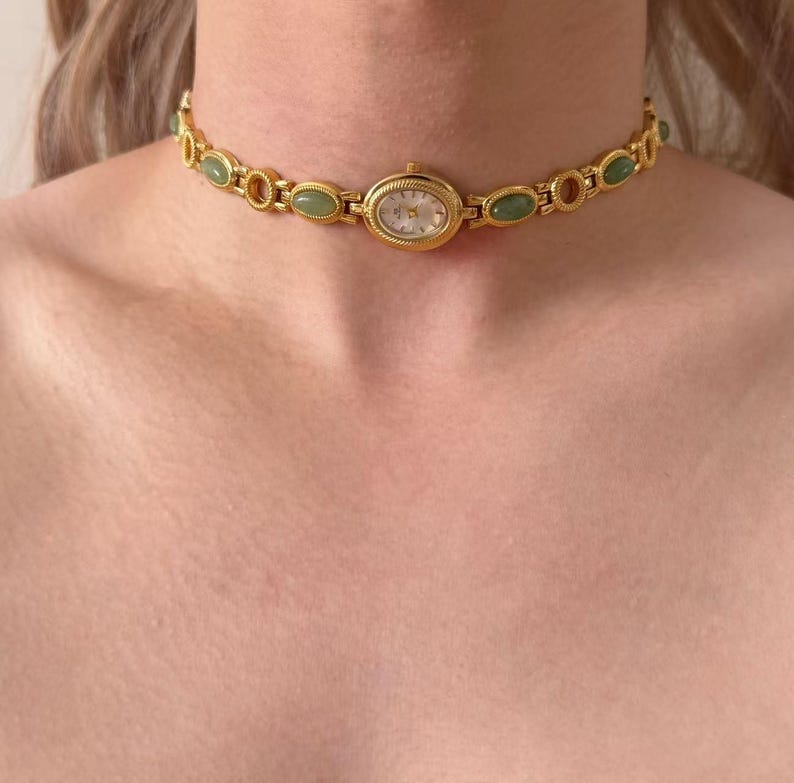 May include: A gold-coloured choker watch featuring an oval face and a white dial. The watch is embellished with green and gold-coloured oval accents. The watch is worn around the neck.