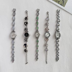 Puede incluir: Cinco relojes de pulsera plateados con esferas ovaladas y correas decorativas. Las correas presentan diversos adornos, incluyendo piedras preciosas, perlas y diseños florales. Las esferas tienen números romanos.