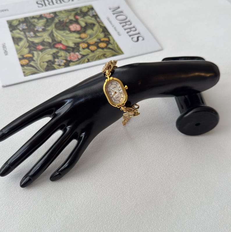 Pu&ograve; includere: Un orologio da polso dorato con quadrante ovale e delicato bracciale a catena. L'orologio presenta un quadrante colorato chiaro con numeri e lancette nere. L'orologio &egrave; esposto su un espositore nero a forma di mano.