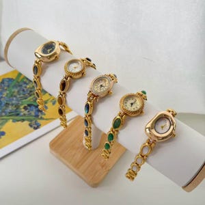 May include: Five gold-toned wristwatches with various oval and round faces are displayed. Each watch has a unique band adorned with colorful gemstones. The watches are presented on a white cylindrical display stand, showcasing their elegant design.