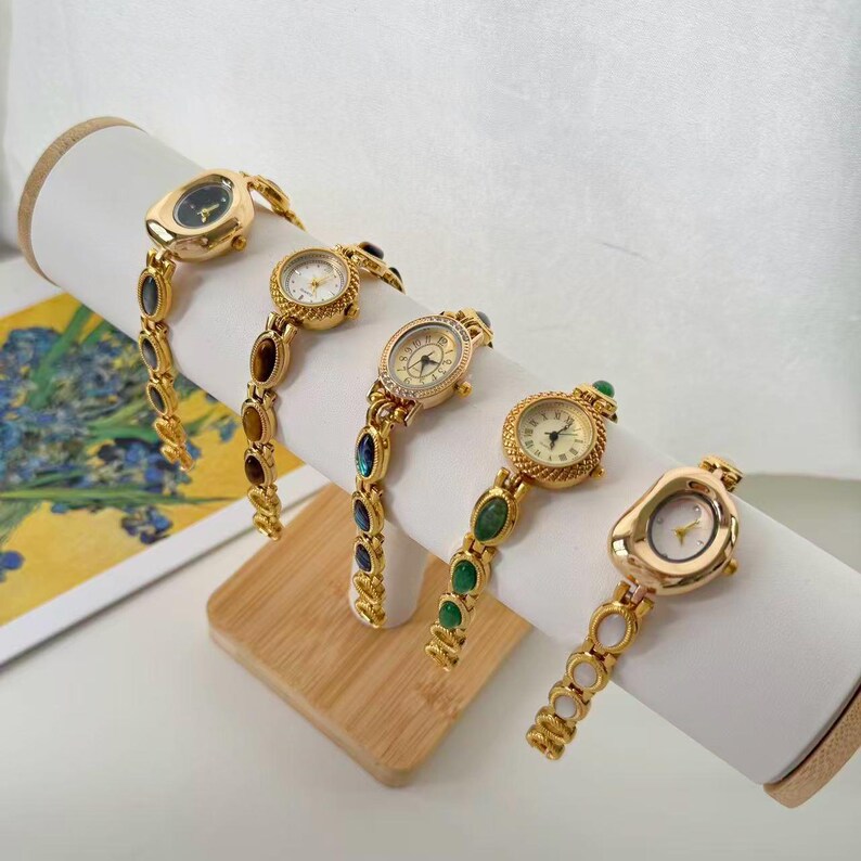 May include: Five gold-toned wristwatches with various oval and round faces are displayed. Each watch has a unique band adorned with colorful gemstones. The watches are presented on a white cylindrical display stand, showcasing their elegant design.