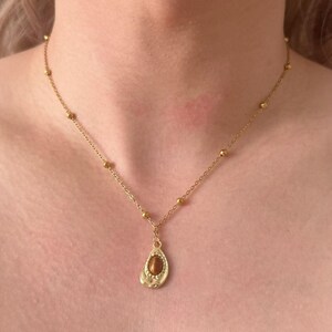 May include: Gold-coloured necklace with a teardrop pendant and a brown stone. The chain has small, round beads. The pendant is approximately 2.5 cm long.