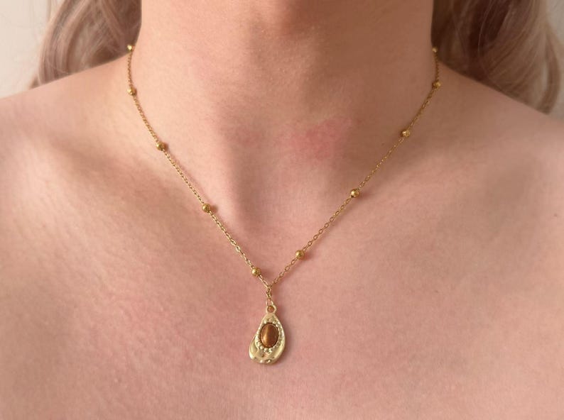 May include: Gold-coloured necklace with a teardrop pendant and a brown stone. The chain has small, round beads. The pendant is approximately 2.5 cm long.