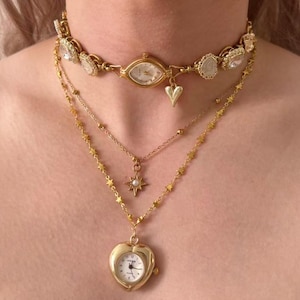 May include: A layered gold-toned necklace set. The top layer is a watch-style choker with clear stones and a heart charm. The second layer has a star pendant with a pearl. The bottom layer features a heart-shaped watch pendant.
