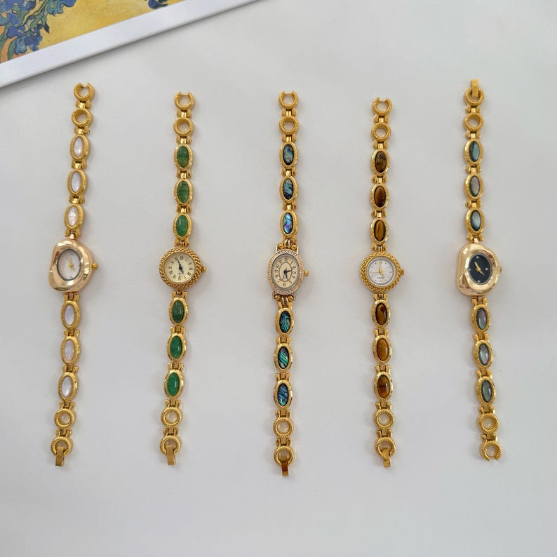 May include: Five gold-toned wristwatches with decorative bands and small watch faces. The bands feature a series of oval-shaped gemstones in various colors, including green, brown, and blue. The watch faces have different designs.