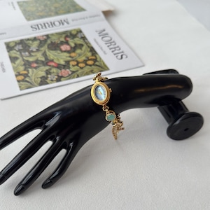 Pu&ograve; includere: Un orologio da polso dorato con quadrante azzurro e dettagli decorativi. L'orologio &egrave; esposto su un espositore nero a forma di mano. L'orologio ha una delicata catena con pietre turchesi e un piccolo ciondolo.