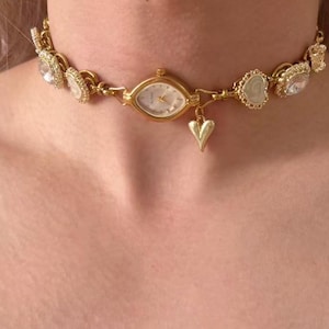 May include: Gold-toned choker necklace featuring a small watch face at the center, surrounded by decorative elements. The necklace includes heart-shaped and gem-like accents. A small heart charm dangles below the watch.