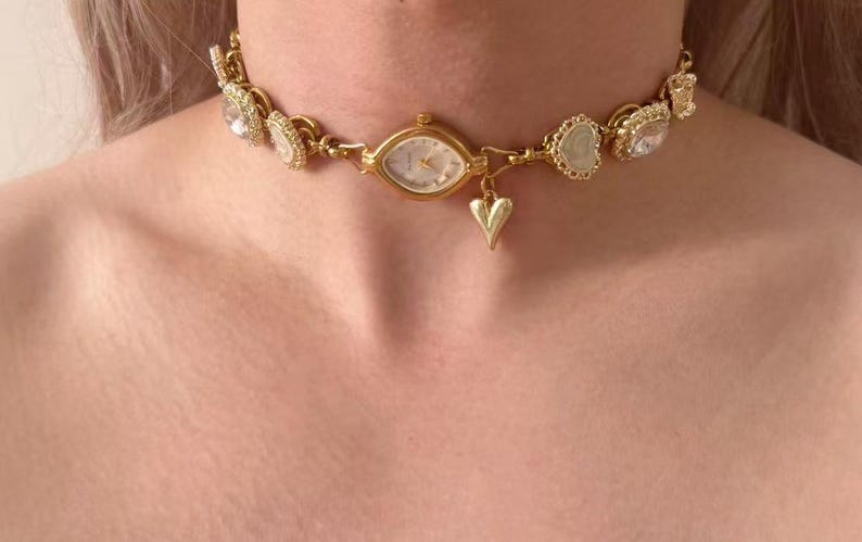 May include: Gold-toned choker necklace featuring a small watch face at the center, surrounded by decorative elements. The necklace includes heart-shaped and gem-like accents. A small heart charm dangles below the watch.