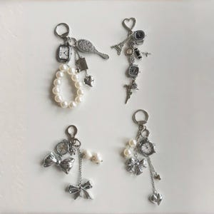 Peut inclure: Quatre porte-clés argentés avec divers breloques, dont des montres miniatures, des nœuds, des perles, des cœurs et une petite Tour Eiffel. Les porte-clés sont disposés sur une surface blanche, mettant en valeur une esthétique délicate et féminine.