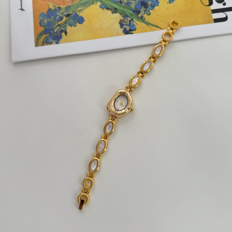 May include: A gold-toned wristwatch with an oval face and a delicate chain. The watch face is white with gold hands and markers. The chain features oval, iridescent stones. A stylish accessory.