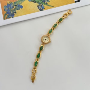 May include: Gold-toned wristwatch with a cream-colored face and black hands. The watch band features oval-shaped green stones set in gold links. The watch is on a white surface.
