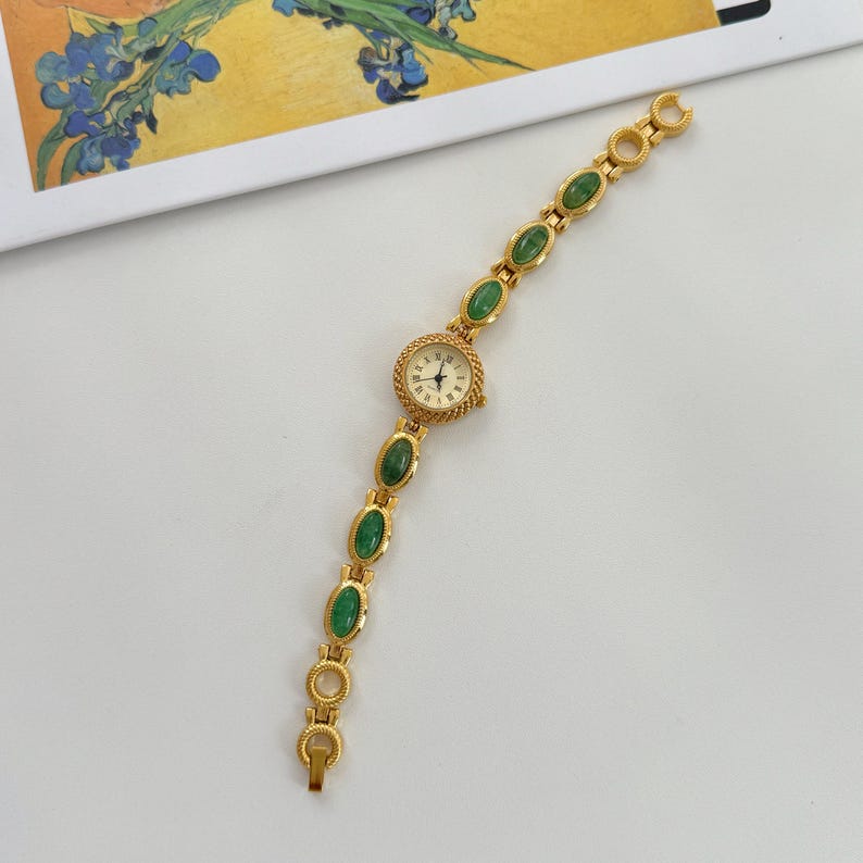 May include: Gold-toned wristwatch with a cream-colored face and black hands. The watch band features oval-shaped green stones set in gold links. The watch is on a white surface.