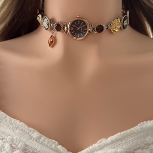 Puede incluir: Un collar gargantilla dorado con una esfera de reloj con esfera marrón oscuro. El collar está adornado con elementos decorativos marrones y dorados, incluido un dije colgante de color ámbar. El reloj tiene un bisel de color oro rosa.