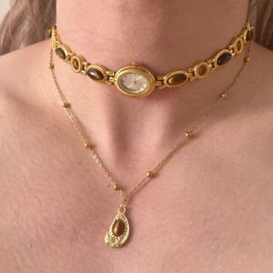 May include: A gold-coloured watch choker and a delicate gold necklace with a teardrop pendant. The watch choker features oval-shaped brown stones and a small watch face. The necklace has a small gold chain with a teardrop-shaped pendant.