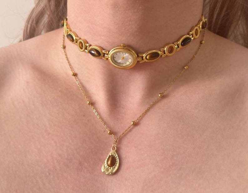 May include: A gold-coloured watch choker and a delicate gold necklace with a teardrop pendant. The watch choker features oval-shaped brown stones and a small watch face. The necklace has a small gold chain with a teardrop-shaped pendant.