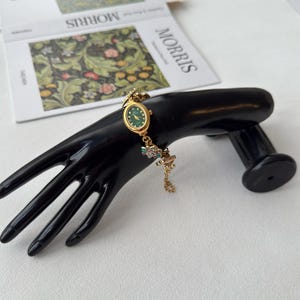 Pu&ograve; includere: Orologio da polso dorato con quadrante verde e delicati dettagli floreali. L'orologio &egrave; esposto su un espositore nero a forma di mano. Il cinturino dell'orologio presenta piccoli abbellimenti colorati. Un libro con la parola "MORRIS" &egrave; sullo sfondo.