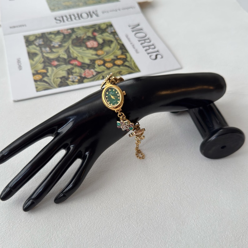 Pu&ograve; includere: Orologio da polso dorato con quadrante verde e delicati dettagli floreali. L'orologio &egrave; esposto su un espositore nero a forma di mano. Il cinturino dell'orologio presenta piccoli abbellimenti colorati. Un libro con la parola "MORRIS" &egrave; sullo sfondo.
