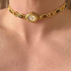 May include: A gold-coloured choker necklace with an oval watch face at the centre. The necklace has oval and circular links with brown-toned stones. The watch face has a white background and gold hands and numerals.