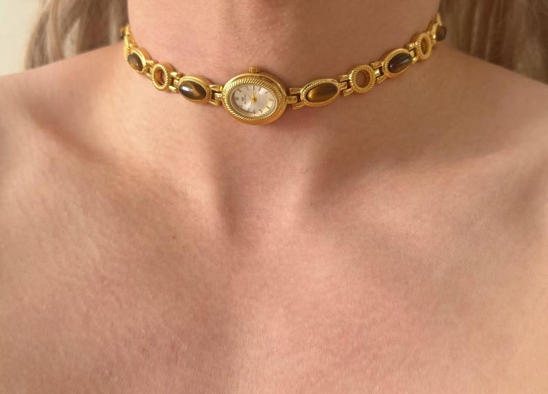 May include: A gold-coloured choker necklace with an oval watch face at the centre. The necklace has oval and circular links with brown-toned stones. The watch face has a white background and gold hands and numerals.